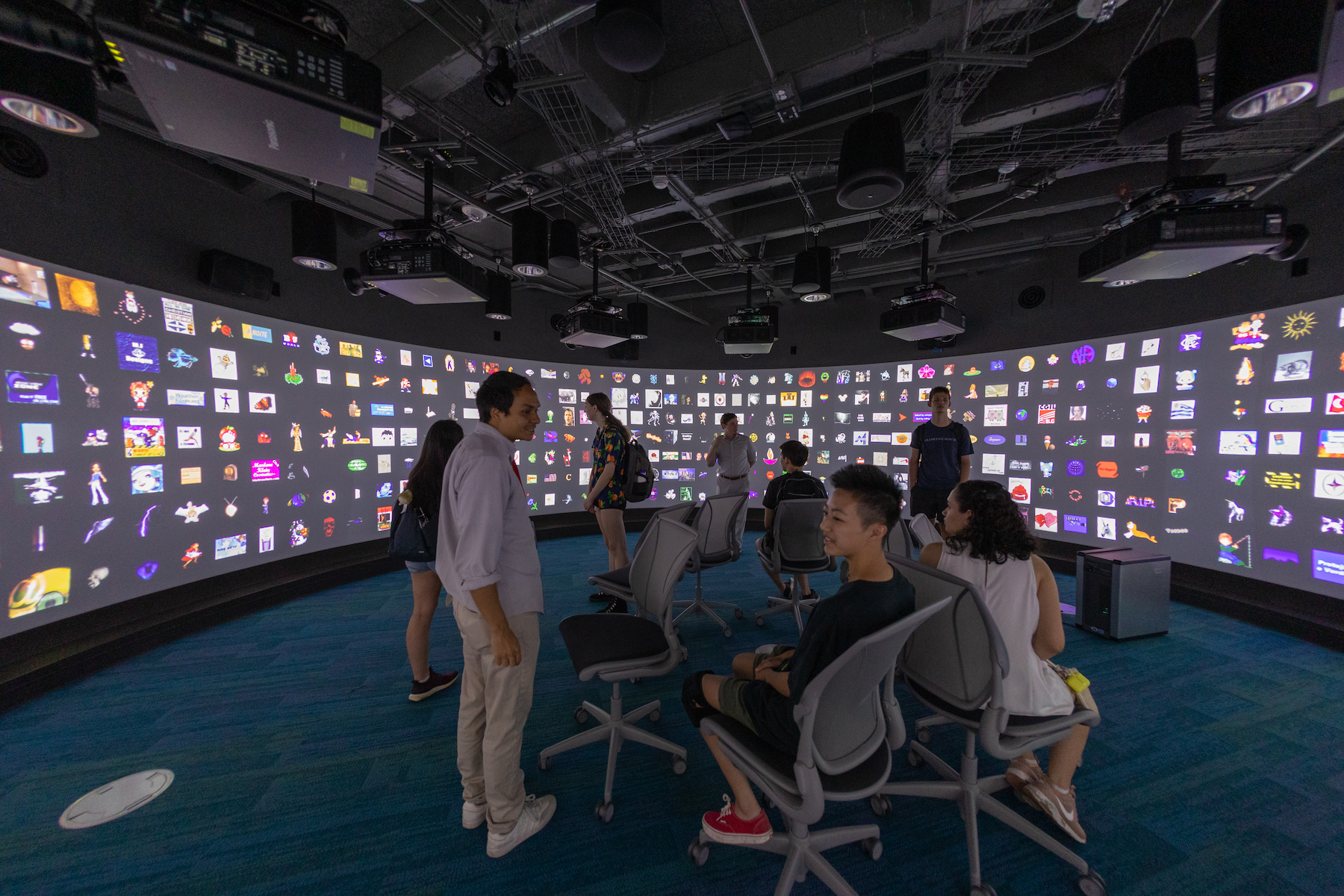 Large curved projection screen with muliple colorful squares. A group of people standing and sitting in the middle of the room. 