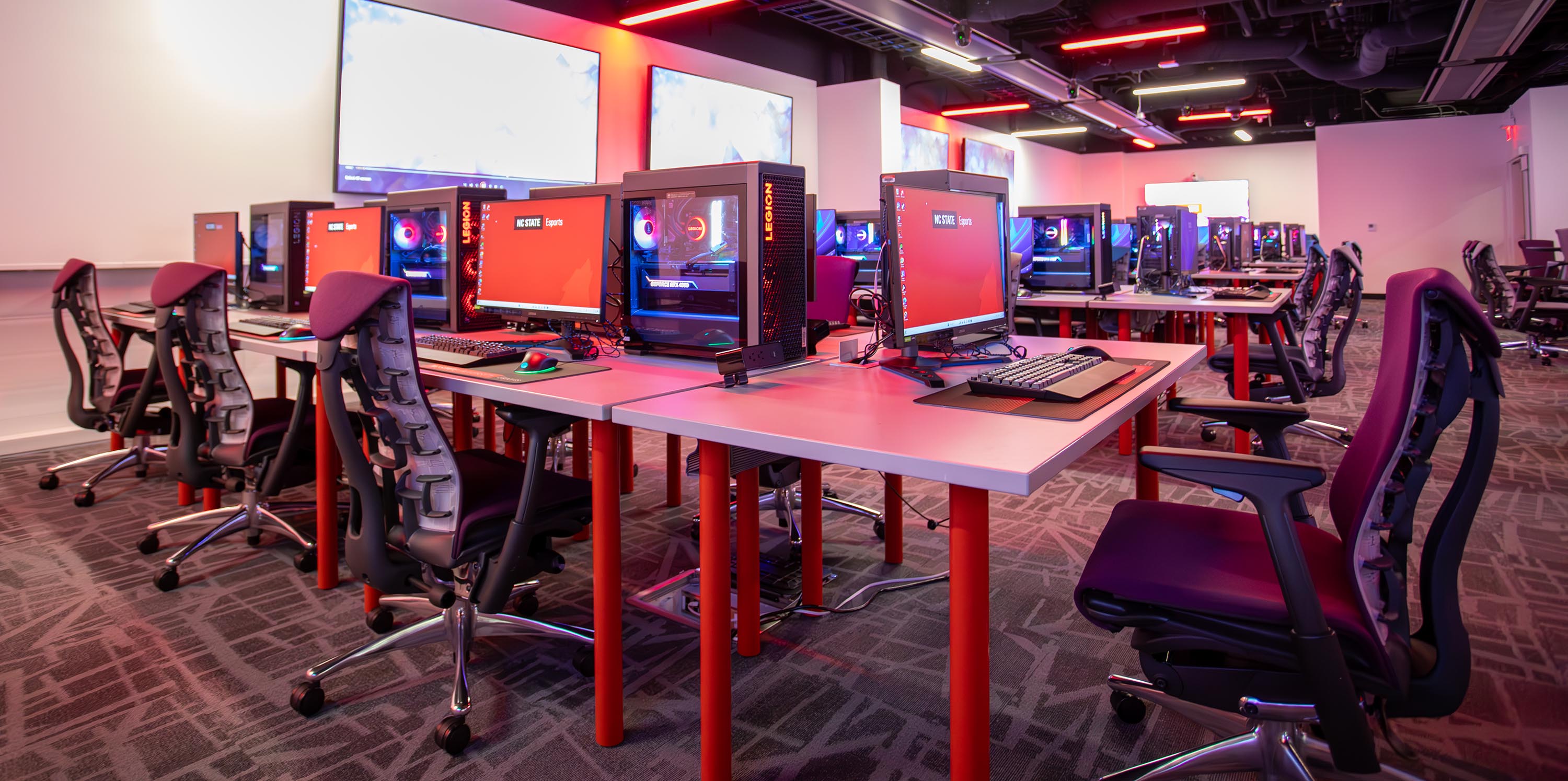 Large room with red lighting, gaming chairs, and tables topped with high-end PCs and monitors