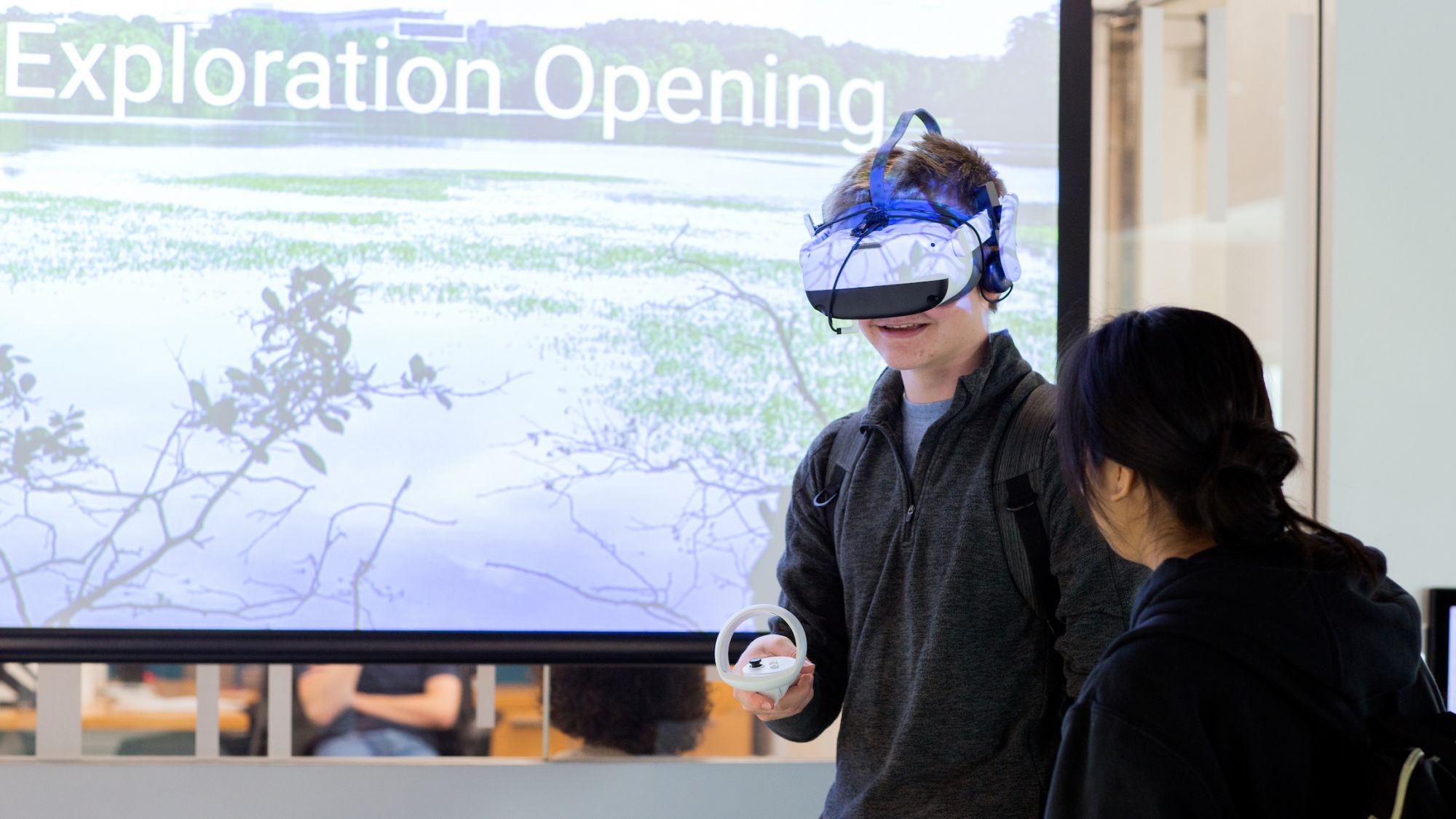 A student wears a virtual reality (VR) headset, with another student standing nearby to assist, in front of a projected image of a natural setting.