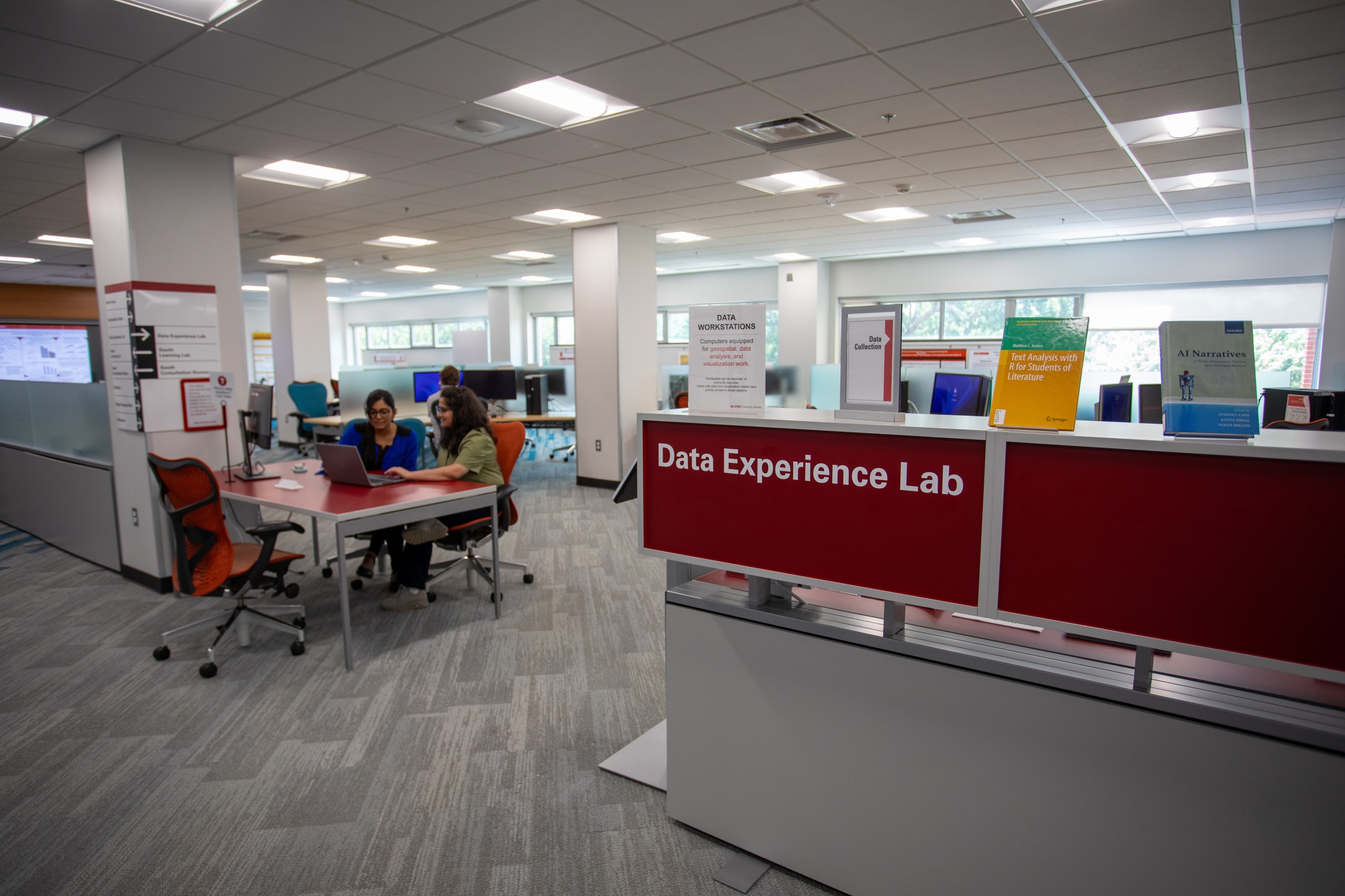 Photo shows a red sign with white lettering indicating the entrance to the "Data Experience Lab," a computing lab with an open and airy design. At the forefront of the image to the left of the sign is a table at which a data science consultant sits with a computer.  She is providing assistance to another student. Behind them, a large wall of windows lights the features of the space. Around the room are computer workstations, a seating area with whiteboard tables, and various data visualizations.