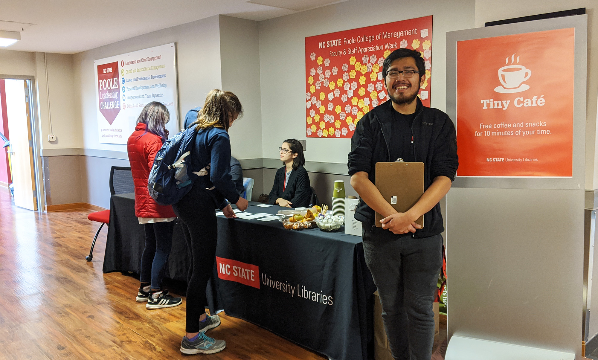 A sign reads, Tiny Café: free coffee and treats for 10 minutes of your time. A smiling student worker with a clipboard is next to it. A long table laden with treats is in the background, with library employees chatting with participants.