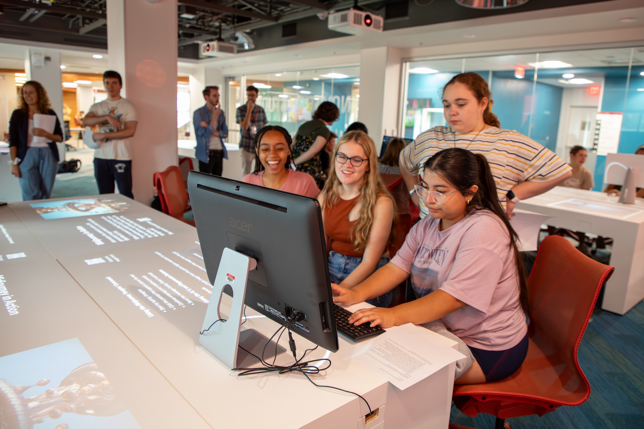 Four students sit at a computer in the Innovation Studio, smiling and laughing. Informative text and images are displayed on the table around the computer.