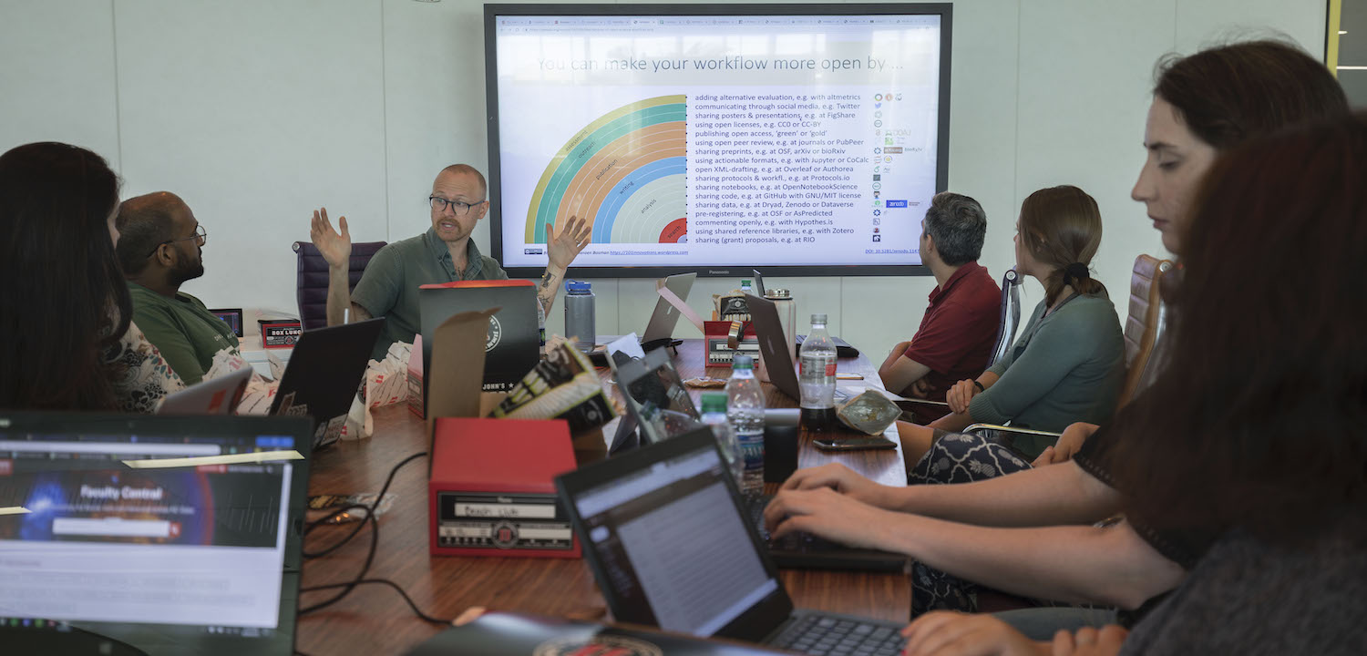 Group of people sitting around a table with laptops discussing a data visualization on a large screen