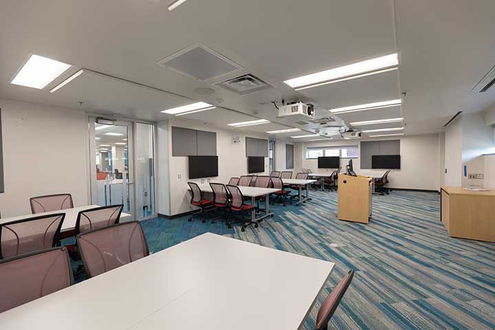 Hill South Learning Lab wide view of room with work tables, chair, digital display screens, ceiling mounted projectors, and podium.