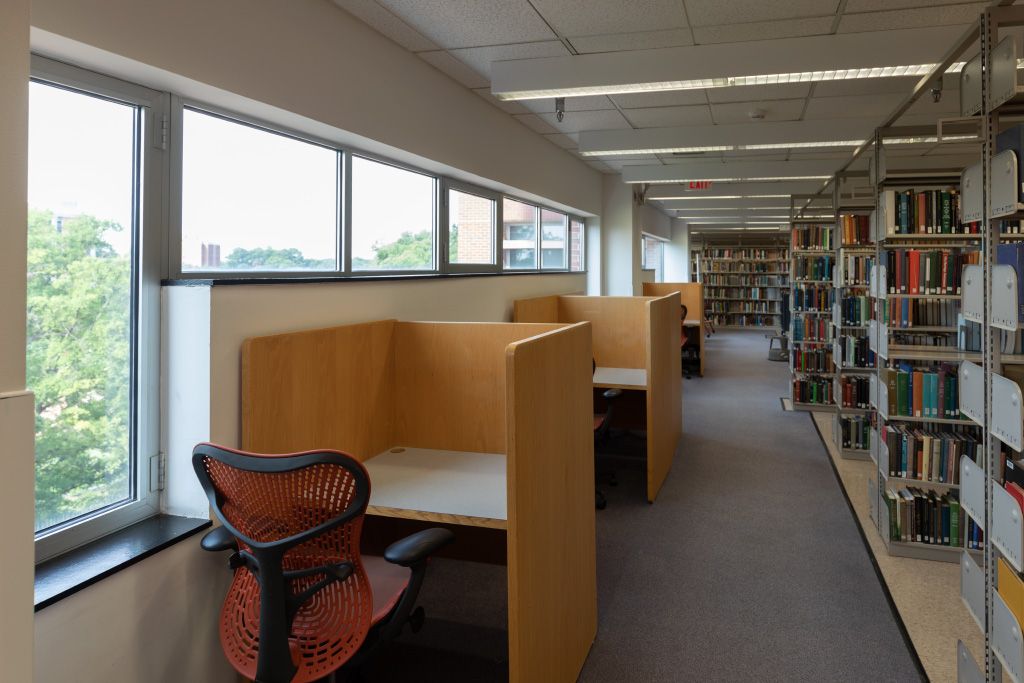 Individual carrels with task chairs along a wall of windows across the aisle from floor to ceiling bookshelves.
