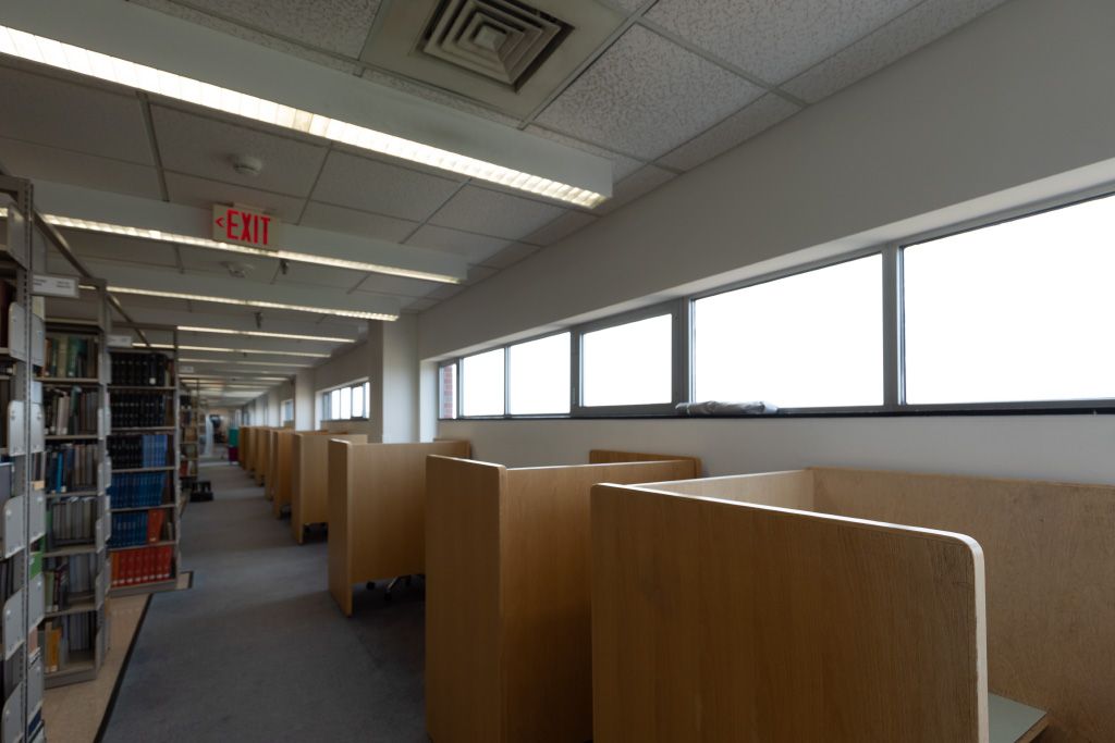 Row of individual carrels with moulded chairs set against a wall with high exterior windows across the aisle from bookshelves.
