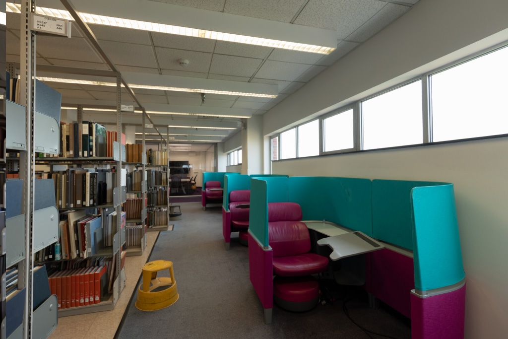 Row of Brody carrels with integrated seat, desk, foot stool, and privacy panels set against a wall with high exterior windows across the aisle from bookshelves.