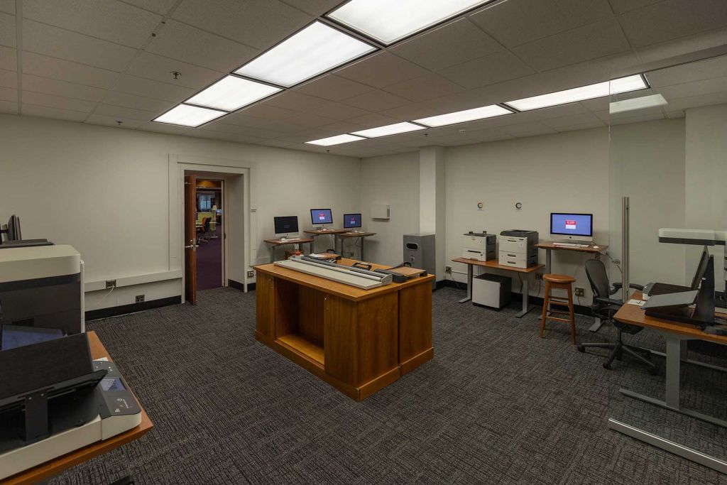 Room with printers, book scanners, and computers on adjustable height desks with central cabinet storage