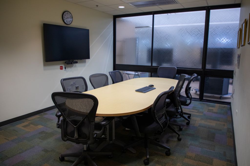 Picture of conference room A109 in the veterinary medicine library. It shows an oval conference table with rolling office chairs and in monitor in the room. The room also features a frosted pane window.