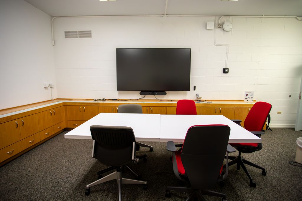 Room with conference table surrounded by 5 chairs and a screen on the wall