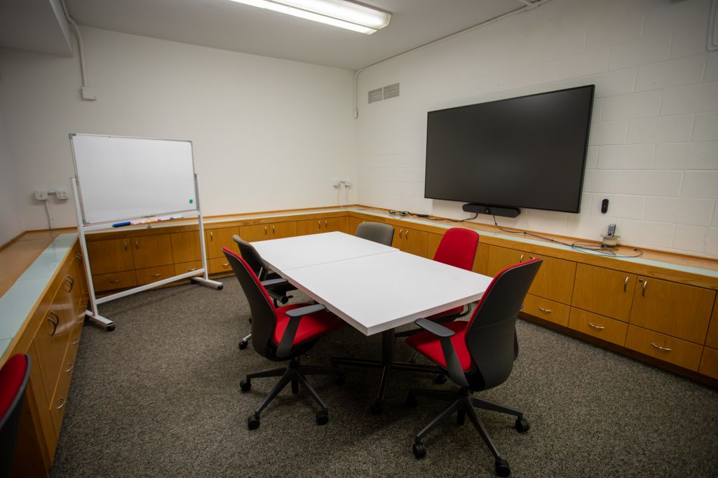 Room with conference table surrounded by 5 chairs, a screen on the wall, and a portable whiteboard