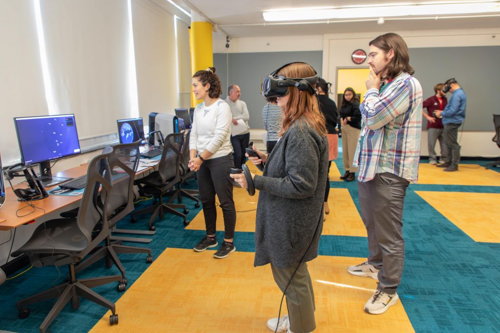 A group of people are participating in a VR training session. A woman in a gray sweater stands in the center, wearing a VR headset and holding controllers, immersed in a virtual environment. A man beside her observes thoughtfully, while a woman in a white sweater looks on, possibly providing guidance.