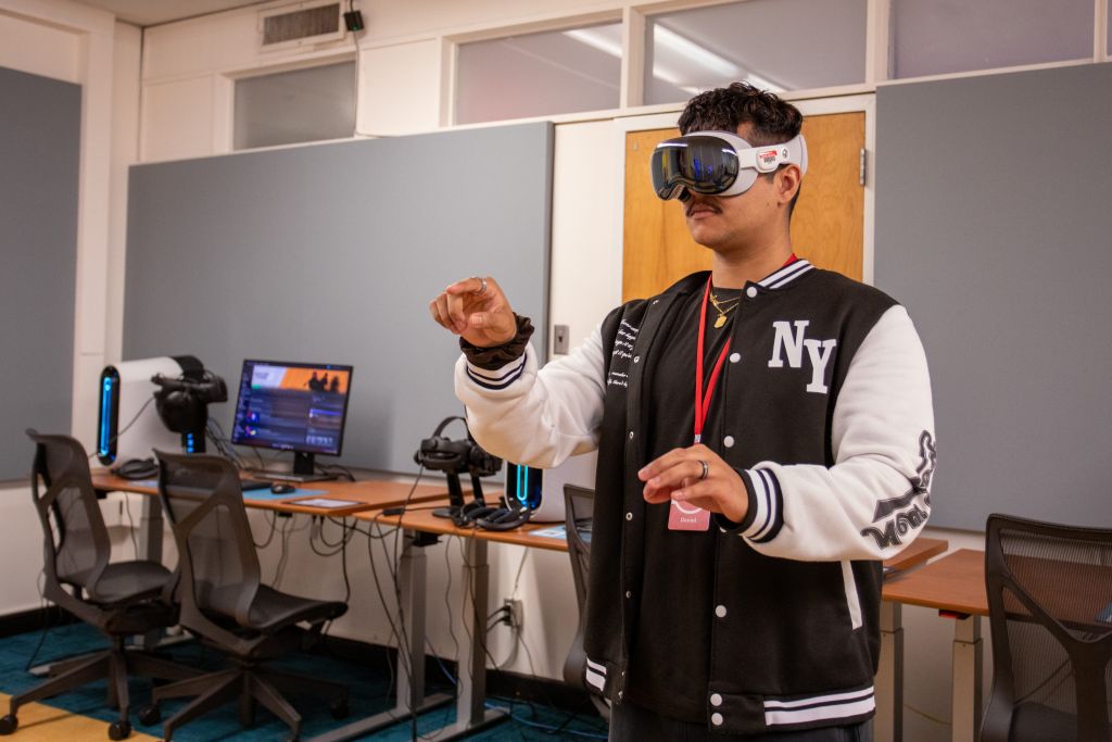  The man is dressed in a black and white jacket and has a name badge. Behind him are several computer workstations equipped with VR headsets and monitors. He's using the Apple Vision Pro spatial computing headset