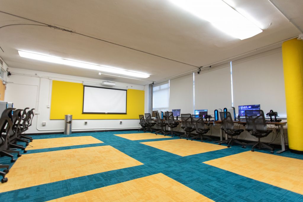 A wide-angle view of the training room, showcasing VR workstations along one side and a large projector screen on the other. The room is well-lit, with modern design elements including colorful carpet tiles and a bright yellow wall. The setup suggests a focus on immersive technology training or workshops.