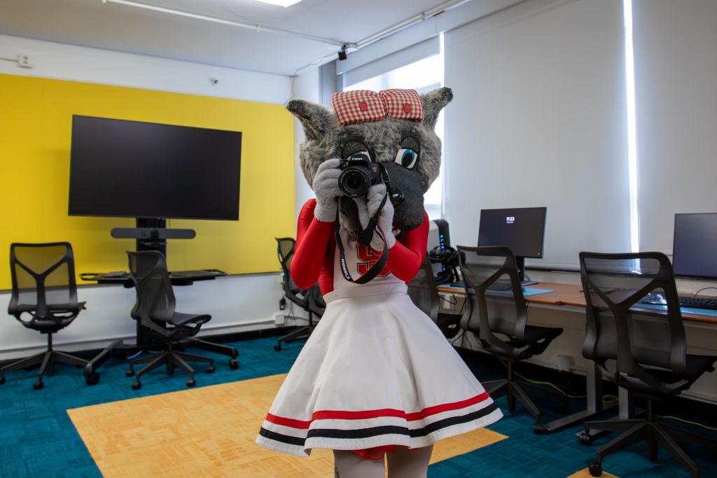 Dressed in a cheerleading outfit with NC State branding, Ms. Wuf holds a DSLR camera, demonstrating the VR studio activities.