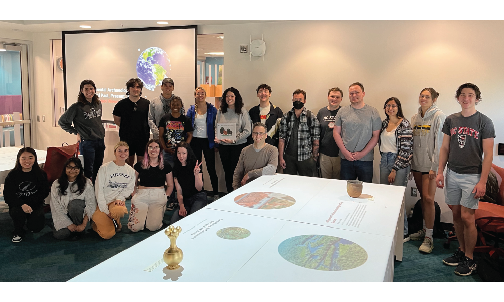 The students of ANT 475 stand around their Innovation Studio exhibit, with digital projections and archaeological artifacts on a white table, with their instructor at center.