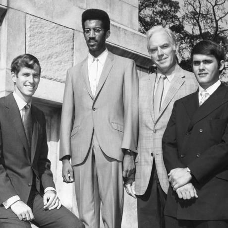 Four people in suits stand in a row, in a black and white photograph