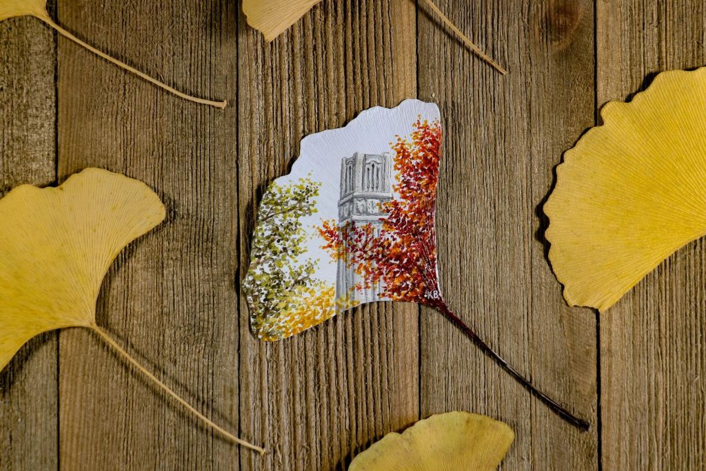 Yellow gingko leaves on wood. One is intricately painted with fall foliage and the NC State Belltower