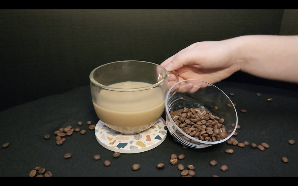 A round glass mug half-full with light brown coffee sits on a colorful terrazo coaster, with a glass bowl of coffee beans aside it, in front of a dark background. A hand reaches in to grab the coffee mug.