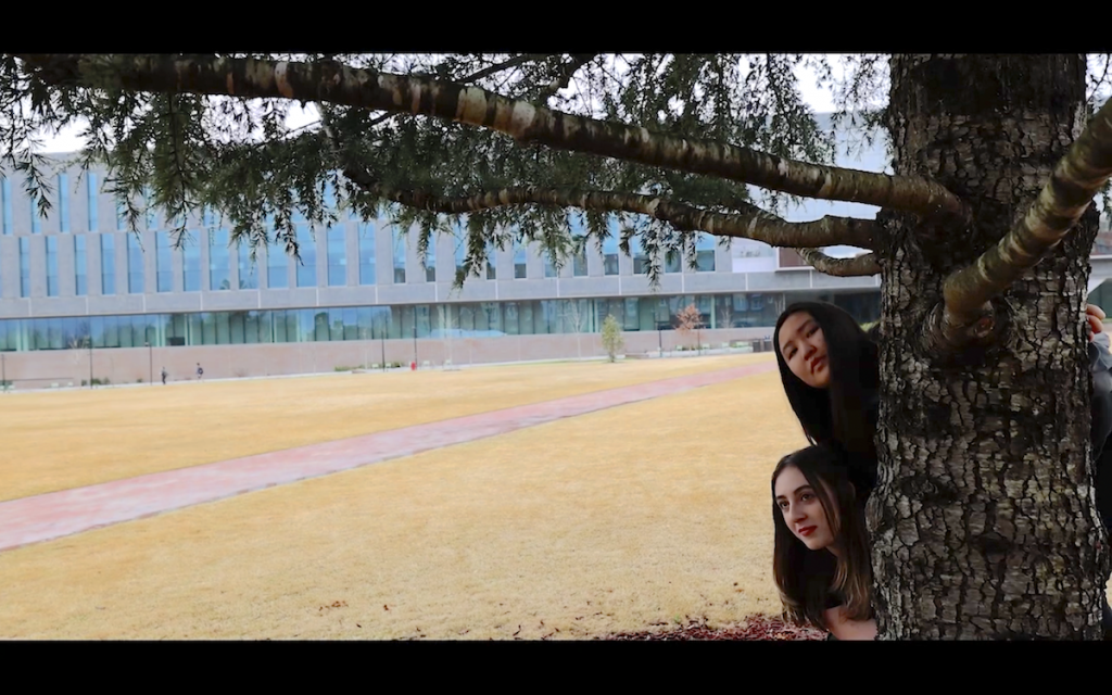 Two students peek their heads out from behind a tree, with a uncertain expressions, hiding at the edge of an open grass square, with a modern college building in the background.