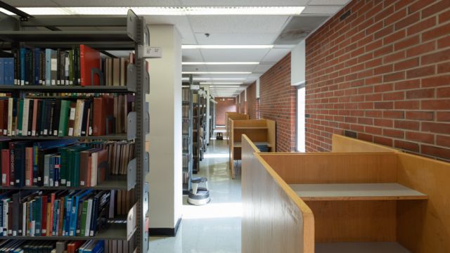 Floor to ceiling bookshelves with books and individual carrel seating along brick wall with exterior windows.