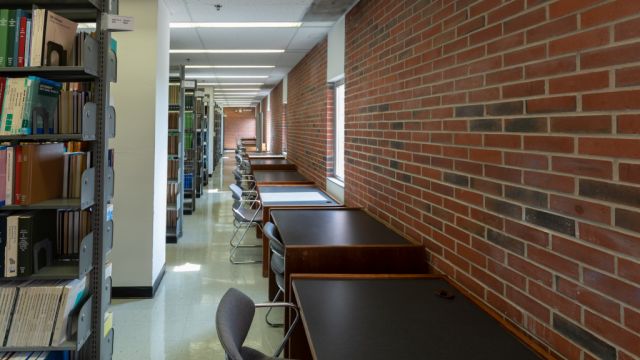 Individual desks with moulded chairs along a brick wall with exterior windows across the aisle from floor to ceiling bookshelves.