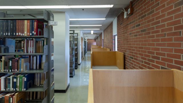 Individual carrels with moulded chairs along a brick wall across the aisle from floor to ceiling bookshelves.