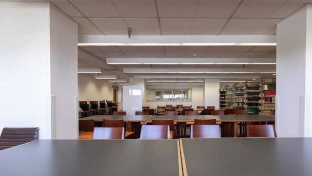 Open space with large tables surrounded by chairs with bookshelves to the right.