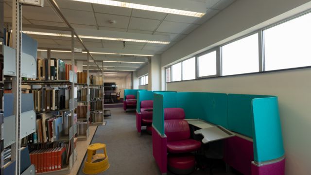 Row of Brody carrels with integrated seat, desk, foot stool, and privacy panels set against a wall with high exterior windows across the aisle from bookshelves.