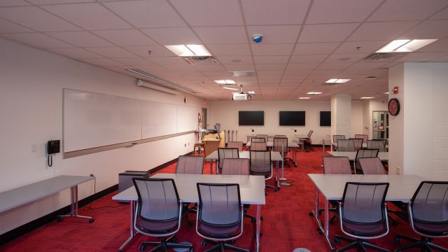 A view from the back of the room showing whiteboards along the left wall and three large screen at the front of the room.