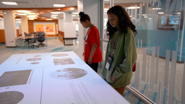 Two students view the exhibits projected onto a table in the Innovation Studio. The projections include text, image, and video and are organized into four stations around the rectangular table.