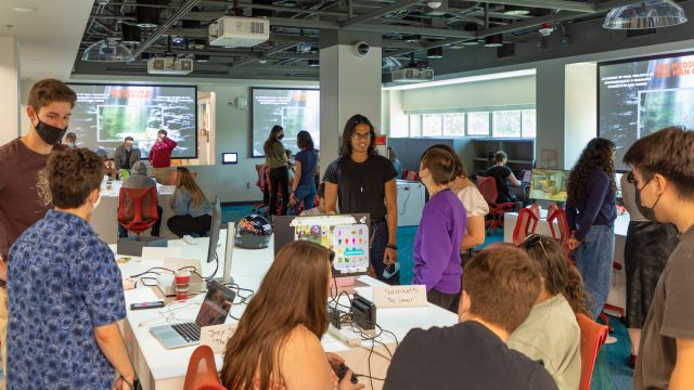 Students gather for a special event in the Innovation Studio, showing their work on tables and viewing each other's projects. There are laptop computers and gaming consoles with screens, as well as physical props.
