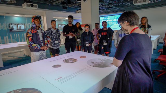 A library staff member introduces the Innovation Studio and its exhibits to a group of students visiting from the Emerging Scholars Academy.