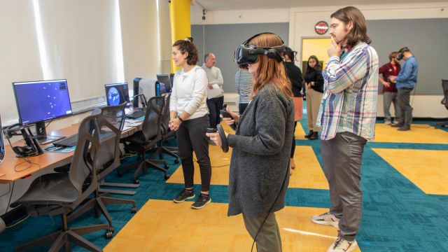 A group of people are participating in a VR training session. A woman in a gray sweater stands in the center, wearing a VR headset and holding controllers, immersed in a virtual environment. A man beside her observes thoughtfully, while a woman in a white sweater looks on, possibly providing guidance.