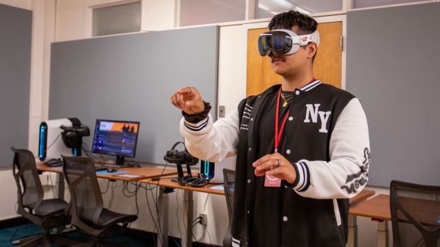  The man is dressed in a black and white jacket and has a name badge. Behind him are several computer workstations equipped with VR headsets and monitors. He's using the Apple Vision Pro spatial computing headset