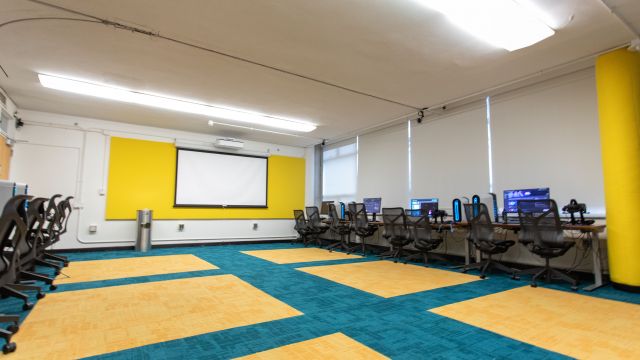 A wide-angle view of the training room, showcasing VR workstations along one side and a large projector screen on the other. The room is well-lit, with modern design elements including colorful carpet tiles and a bright yellow wall. The setup suggests a focus on immersive technology training or workshops.