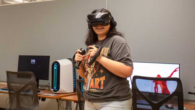 A participant is engaged in a virtual reality experience in a well-equipped room. She stands in front of a desk setup with a high-performance desktop PC and a monitor displaying a red abstract figure. The room features modern design elements with ergonomic chairs and clean lines.