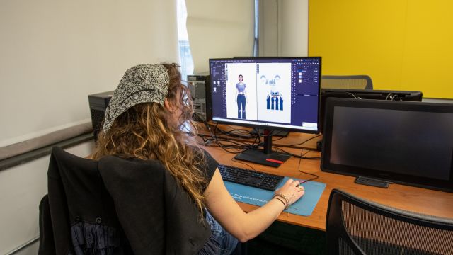 The artist is working on a dual-monitor setup. The screen shows a digital illustration of a character with various poses and elements, indicating work on character design or animation.