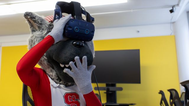Ms Wuf wears a VR headset and is surprised with the virtual environment. The mascot's expression suggests excitement and engagement.