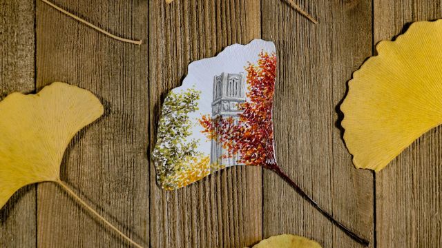 Yellow gingko leaves on wood. One is intricately painted with fall foliage and the NC State Belltower