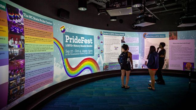 A wide screen displays a colorful exhibit with the title, PrideFest, LGBTQ+ History Month Exhibit