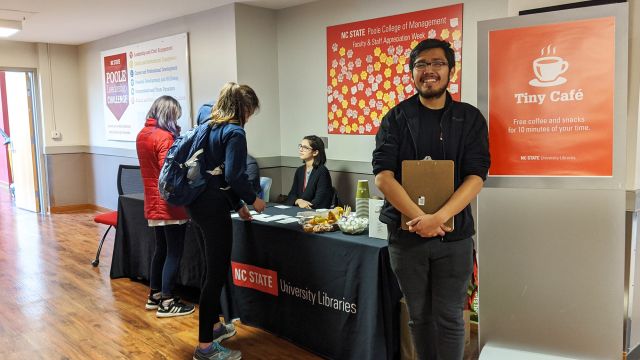 A sign reads, Tiny Café: free coffee and treats for 10 minutes of your time. A smiling student worker with a clipboard is next to it. A long table laden with treats is in the background, with library employees chatting with participants.