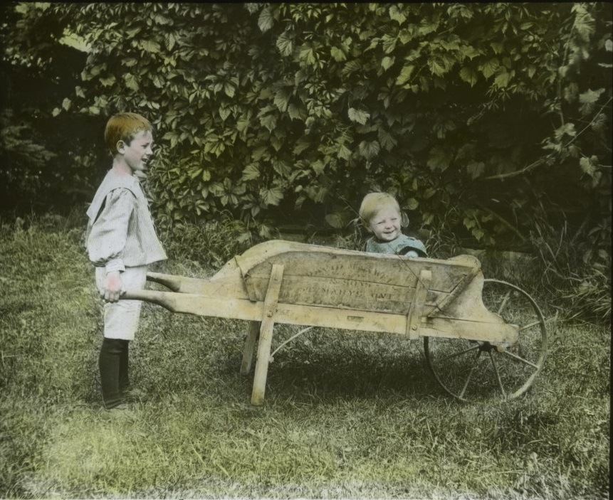 Young boy wheeling younger boy in wheelbarrow, colorized, circa 1910